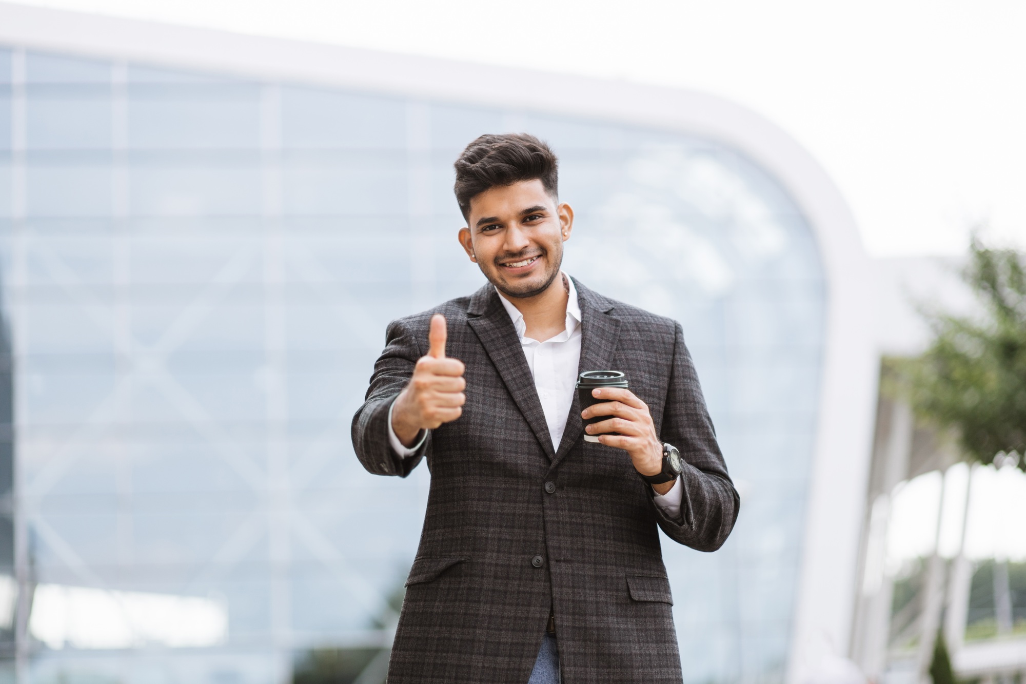 Arab businessman holding a cup of coffee looking on camera and showing thumb up approval gesture.